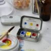 open tin of Handmade watercolour paint by Art Scribe Primary Collection. A paintbrush, palette with watercolour paint and paper with splashes of colour from the tin sit on the table. Jars and bottles are in the background.