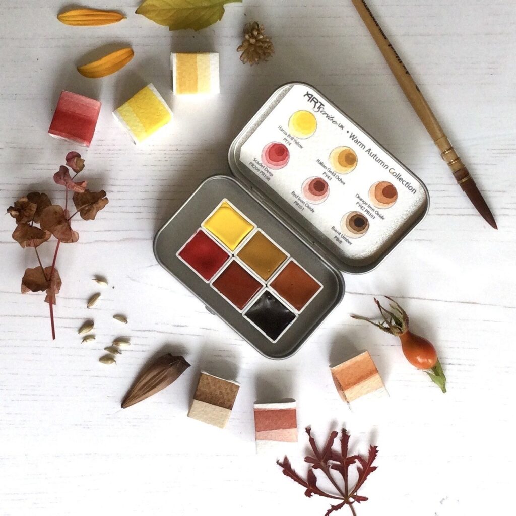 image shows a small tin open tin containing six colours inspired by the autumn season. The yellows, reds and brown colours of the paint are reflected in the natural autumn objects such as nuts and leaves that are scattered around the tin.