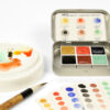 Open tin of Japanese Handmade Watercolour Collection by Art Scribe sits on the table with a paintbrush, paint palette and gradients of the six colours.