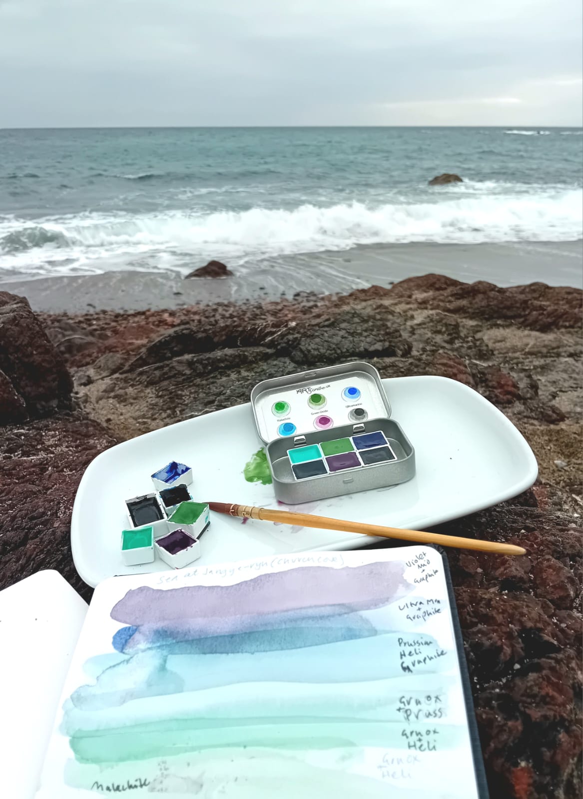 in the foreground lies an open sketchbook with colours used from the Selkies Collection. A mixing palette and open tin of the set sit on a rock that overlooks the sea.