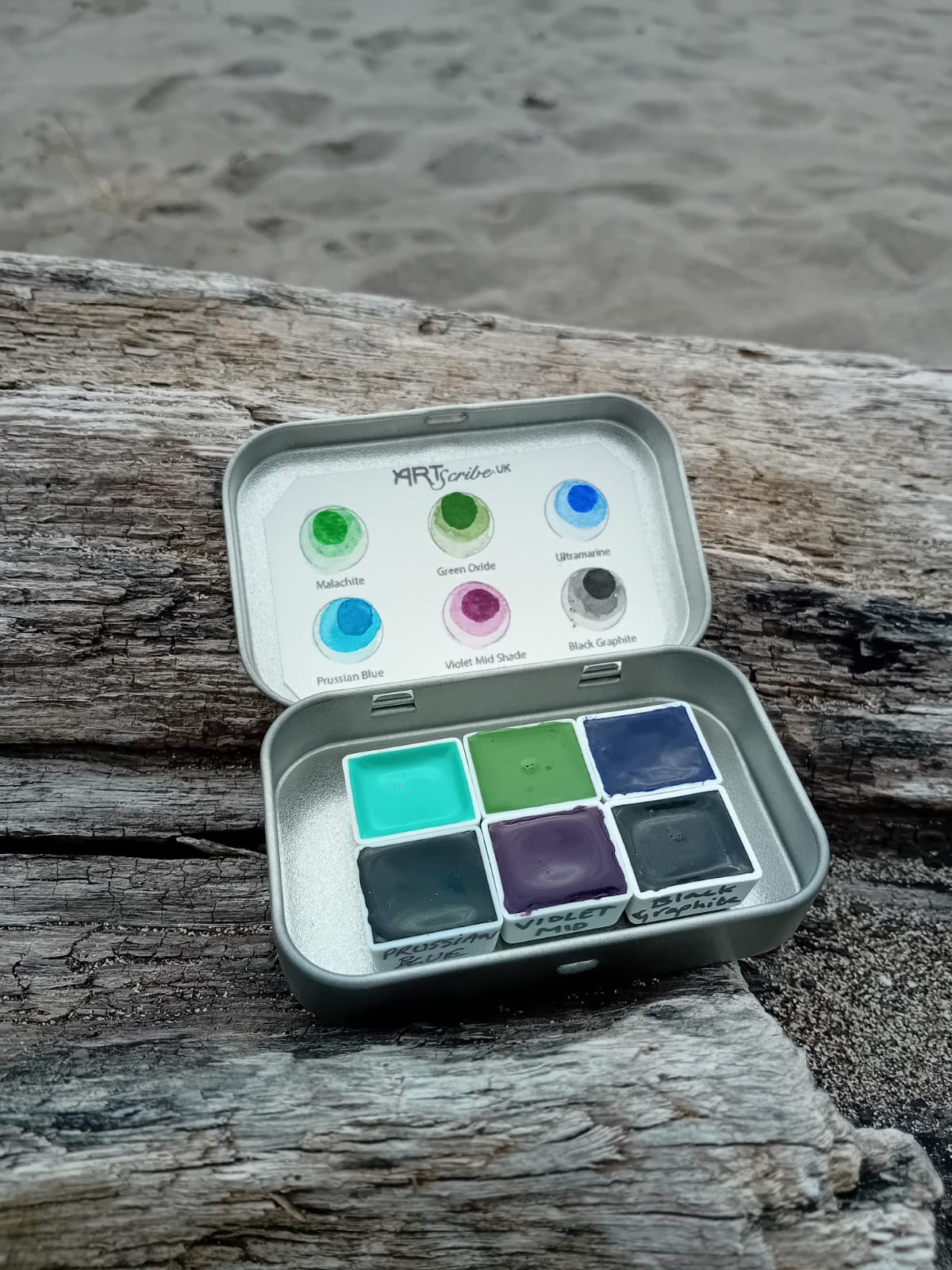 Image shows an open tin of watercolour paints that make up the Selkies Collection. The colours include blues, green and violet. The tin sits on a piece of drift wood.
