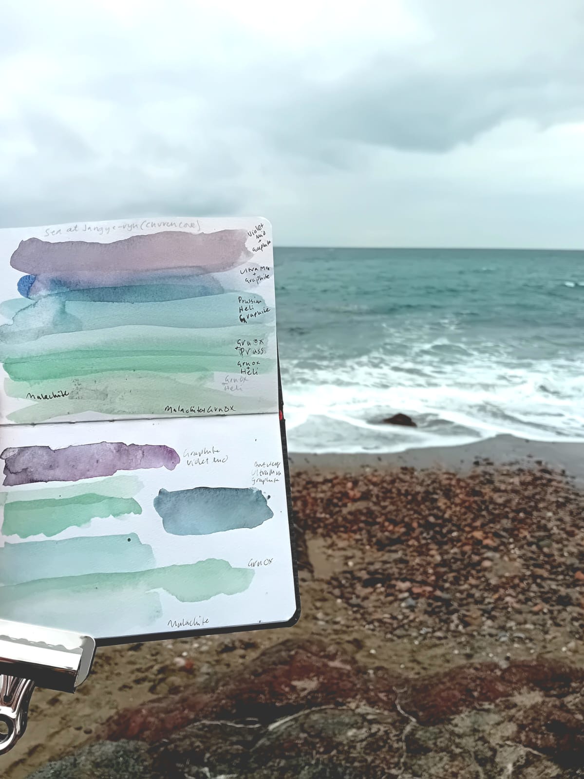 Held up against the background of the sea is an open sketchbook. The pages of the sketchbook contain loose watercolour washes that reflect the colours of the sea.