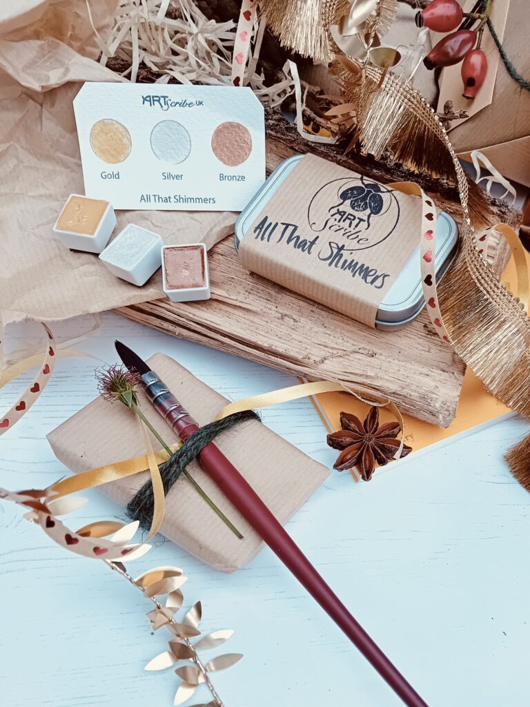 Three pans of watercolour paint in colours gold, silver and bronze, sit next to an Art Scribe tin and paint brush, surrounded by wrapping paper and gold ribbon.