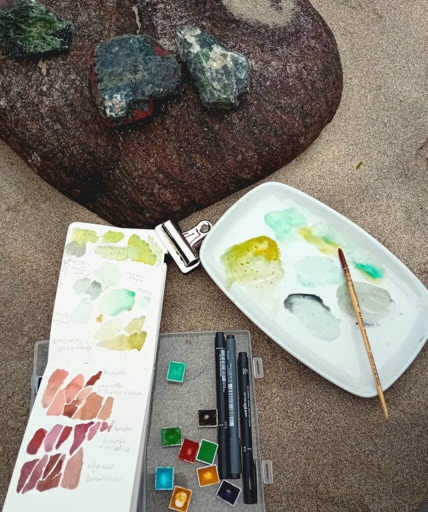 On a sandy beach sits a dark red boulder of serpentine and several smaller rocks of gray and green serpentine. next to the boulder is an open sketchbook with colour notes, several watercolour pans, paint brushes and pens and a mixing palette of greens.